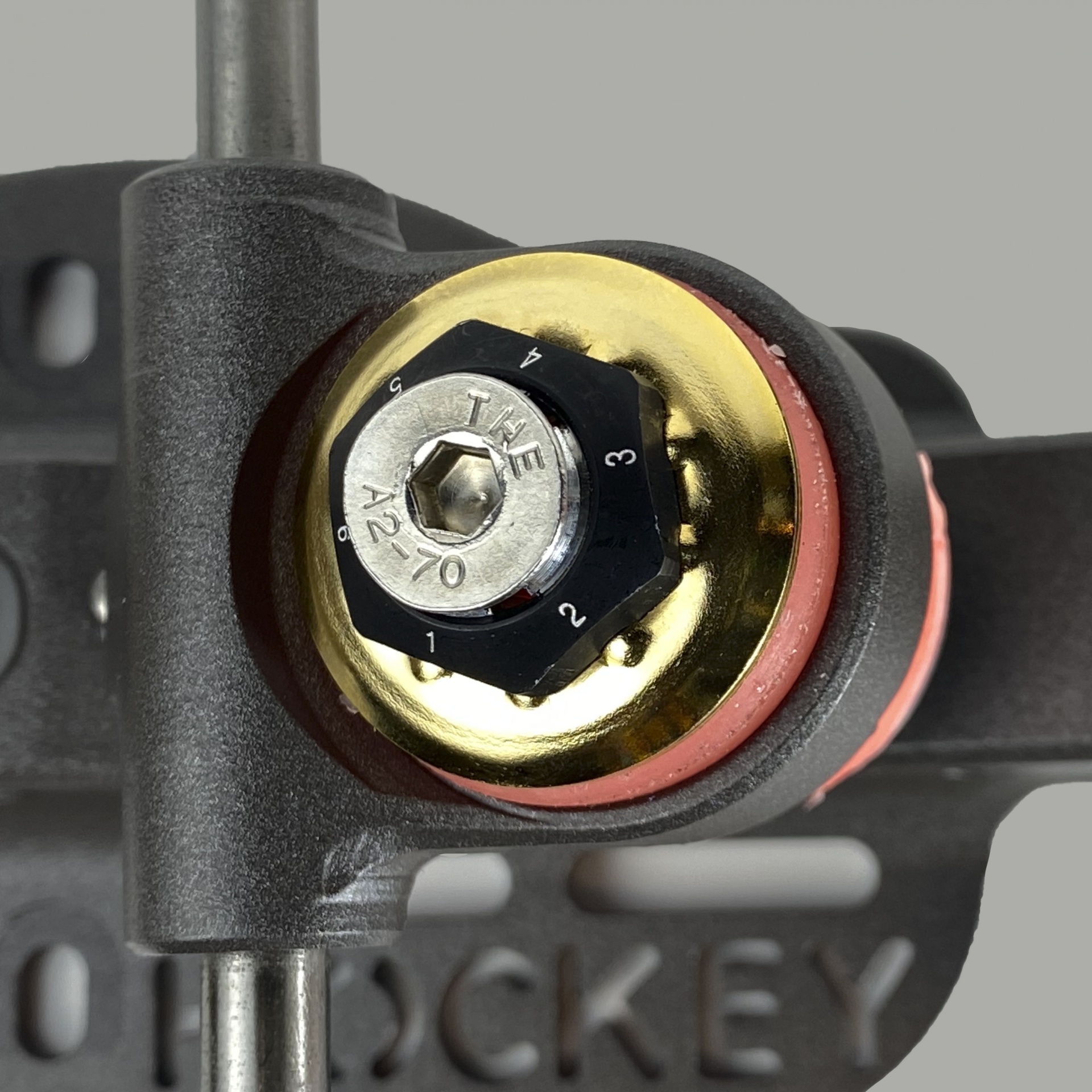 Peça mecânica com porca preta, aro dourado e anel vermelho sobre fundo escuro com texto parcial