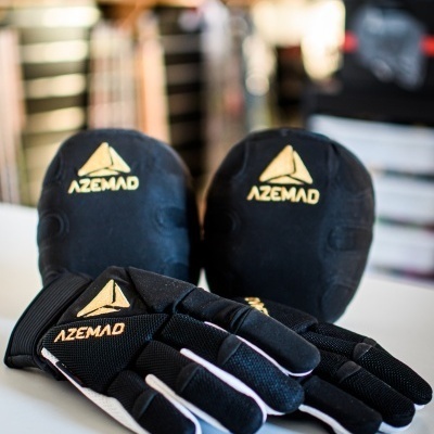 Luvas e joelheiras pretas com logótipo AZEMAD dourado sobre superfície branca