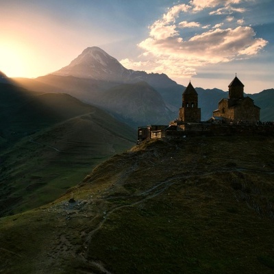 Igreja antiga em colina montanhosa ao pôr do sol com céu nublado e luz solar ao fundo