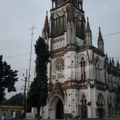 Igreja gótica branca e castanho-avermelhada com torres pontiagudas numa rua com pessoas e árvores