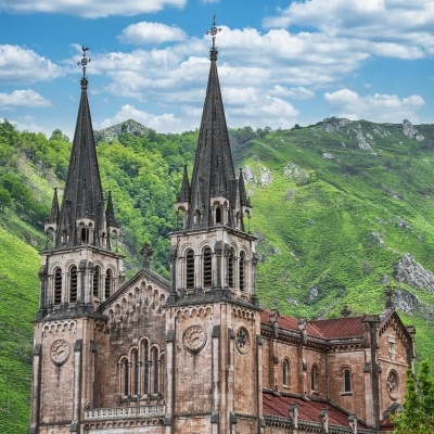Igreja em pedra com duas torres pontiagudas e relógios, rodeada por vegetação e colinas verdes