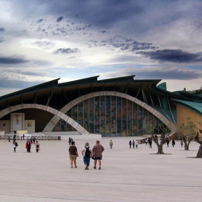 Edifício com telhado curvo e fachada em vidro colorido, espaço pavimentado e árvores sob céu nublado