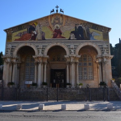 Fachada de igreja com arcos, colunas e mosaicos religiosos coloridos, cercada por rua e árvores