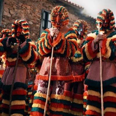 Pessoas com trajes tradicionais coloridos e máscaras segurando paus numa rua de pedra junto a edifício de pedra