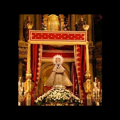 Estátua religiosa feminina com manto dourado em altar com cortinas vermelhas e velas.
