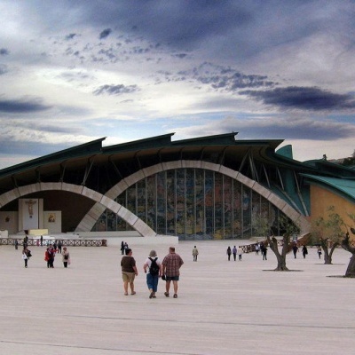 Edifício moderno com fachada em vidro curvo e arcos brancos, cobertura verde, praça ampla com pessoas e árvores