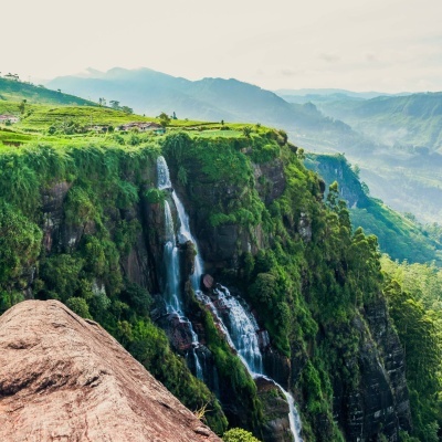 Cascata caindo por falésia verde com montanhas ao fundo