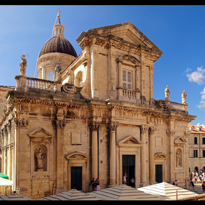 Edifício histórico barroco em pedra clara com cúpula e esculturas, céu azul, zona urbana e pessoas