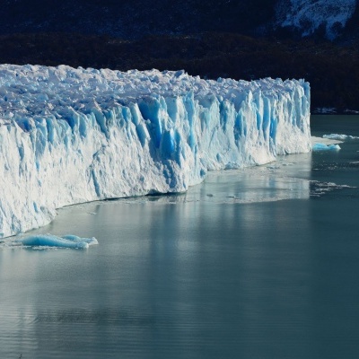 Geleira branca e azul clara à beira de água com montanhas ao fundo