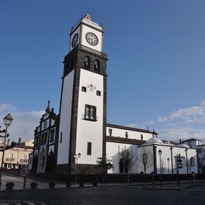 Igreja branca com torre de relógio em praça com pavimento de paralelos