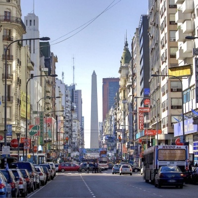 Rua movimentada numa cidade com edifícios altos, carros estacionados e o Obelisco ao fundo
