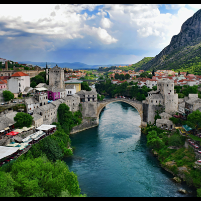 Ponte histórica em arco sobre rio azul turquesa, rodeada por edifícios em pedra e torres, com montanhas ao fundo
