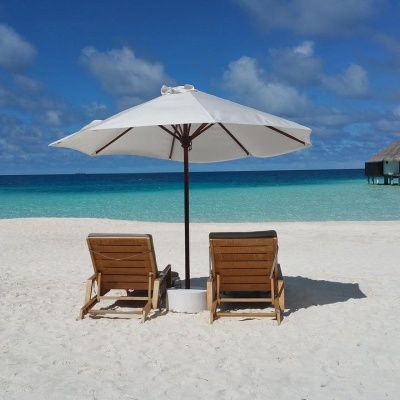 Duas espreguiçadeiras de madeira com guarda-sol branco numa praia com areia branca e mar azul