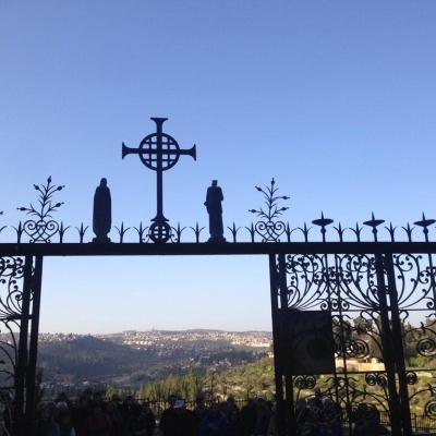 Portão de metal negro com cruz e estátuas, vista de colinas ao fundo