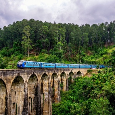 Comboio azul numa ponte de pedra com arcos, em ambiente natural verde e céu nublado