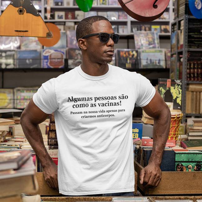 Algumas pessoas são como as vacinas! Passam na nossa vida apenas para criarmos anticorpos. Homem de camiseta branca com texto em português numa loja de discos e livros
