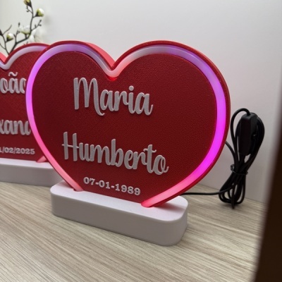 Luminária coração vermelho com luz LED rosa, nomes e data em letras prateadas, sobre mesa clara