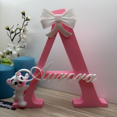 Letra decorativa rosa com texto Aurora e ursinho branco, sobre superfície de madeira com velas azuis e flores artificiais.