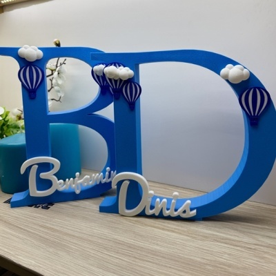 Decoração com letras azuis B e D com nomes Benjamin e Dinis em branco, decoradas com balões e nuvens.