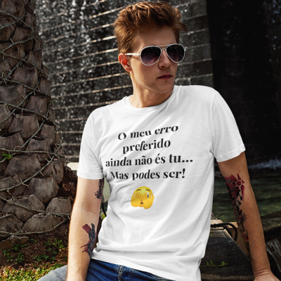 Camiseta branca com texto e emoji usada por jovem com tatuagens e óculos escuros sentado perto de tronco de palmeira