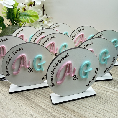 Placas decorativas redondas de madeira com letras A e G em rosa e azul, texto Batiismo Ariel & Gabriel, data 25 Outubro 2026, em suporte branco.