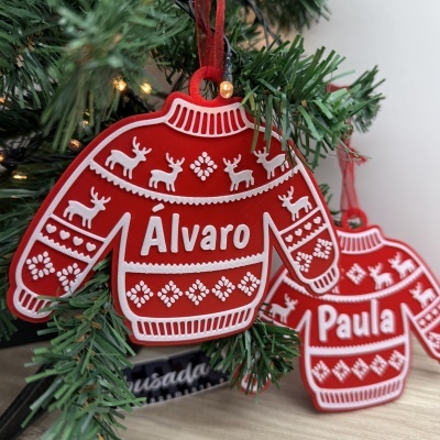 Enfeites de Natal em forma de camisolas vermelhas com padrões brancos e nomes personalizados pendurados numa árvore verde.