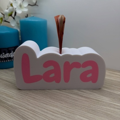 Cofre de espuma branco com nome Lara em letras cor-de-rosa e nota de euro dobrada, velas azuis e flores ao fundo.