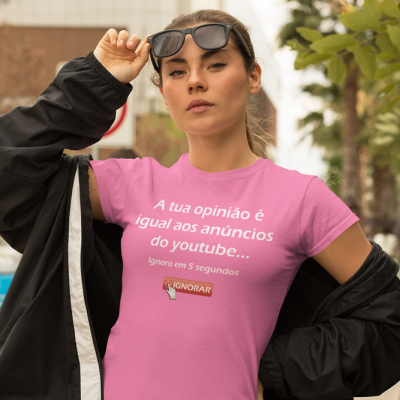 Pessoa jovem com t-shirt rosa com texto e óculos de sol com casaco preto num ambiente urbano.