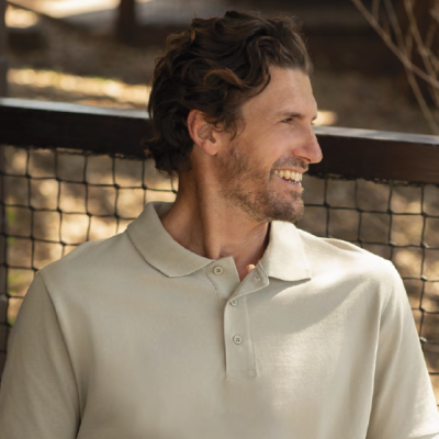 Homem com camisa polo bege sorrindo ao ar livre encostado numa rede metálica