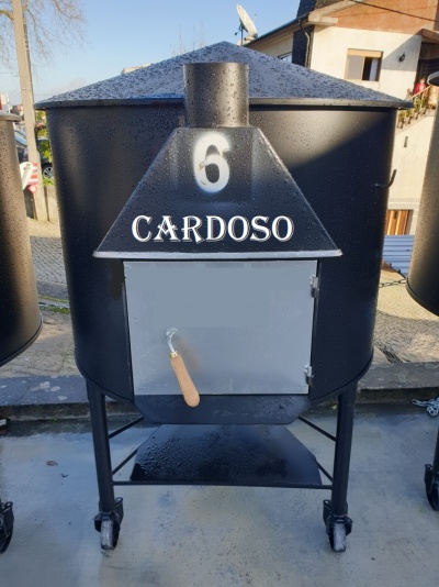 Forno preto com porta cinza e inscrições brancas, sobre rodas em pavimento exterior