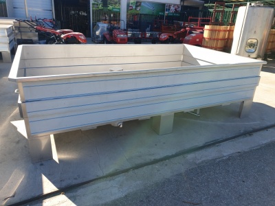 Contentor metálico retangular cinza claro sobre pernas, com máquinas agrícolas vermelhas ao fundo