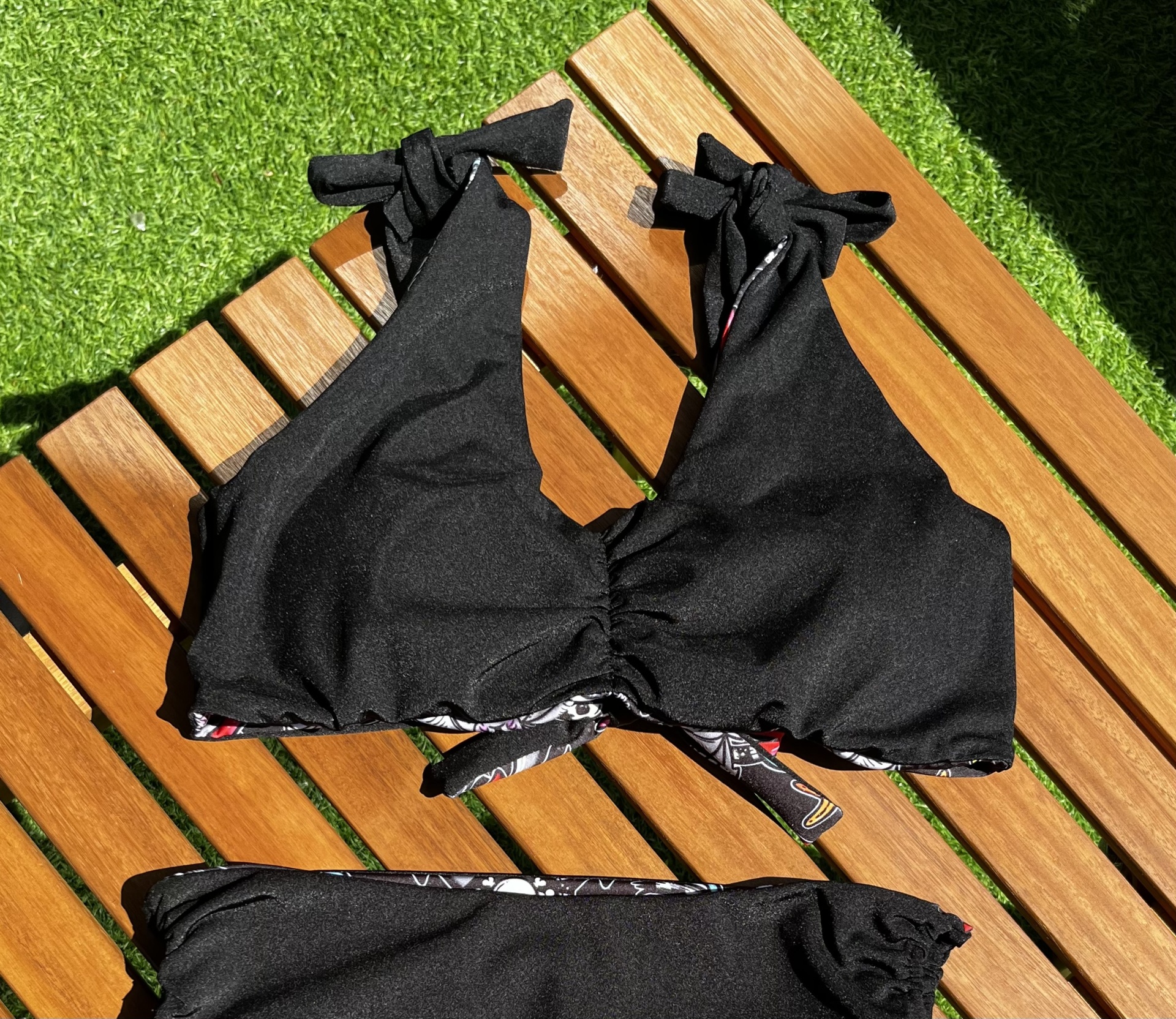 Biquíni preto com alças de laço, tecido liso, parte inferior com padrão floral, sobre madeira clara e relva sintética