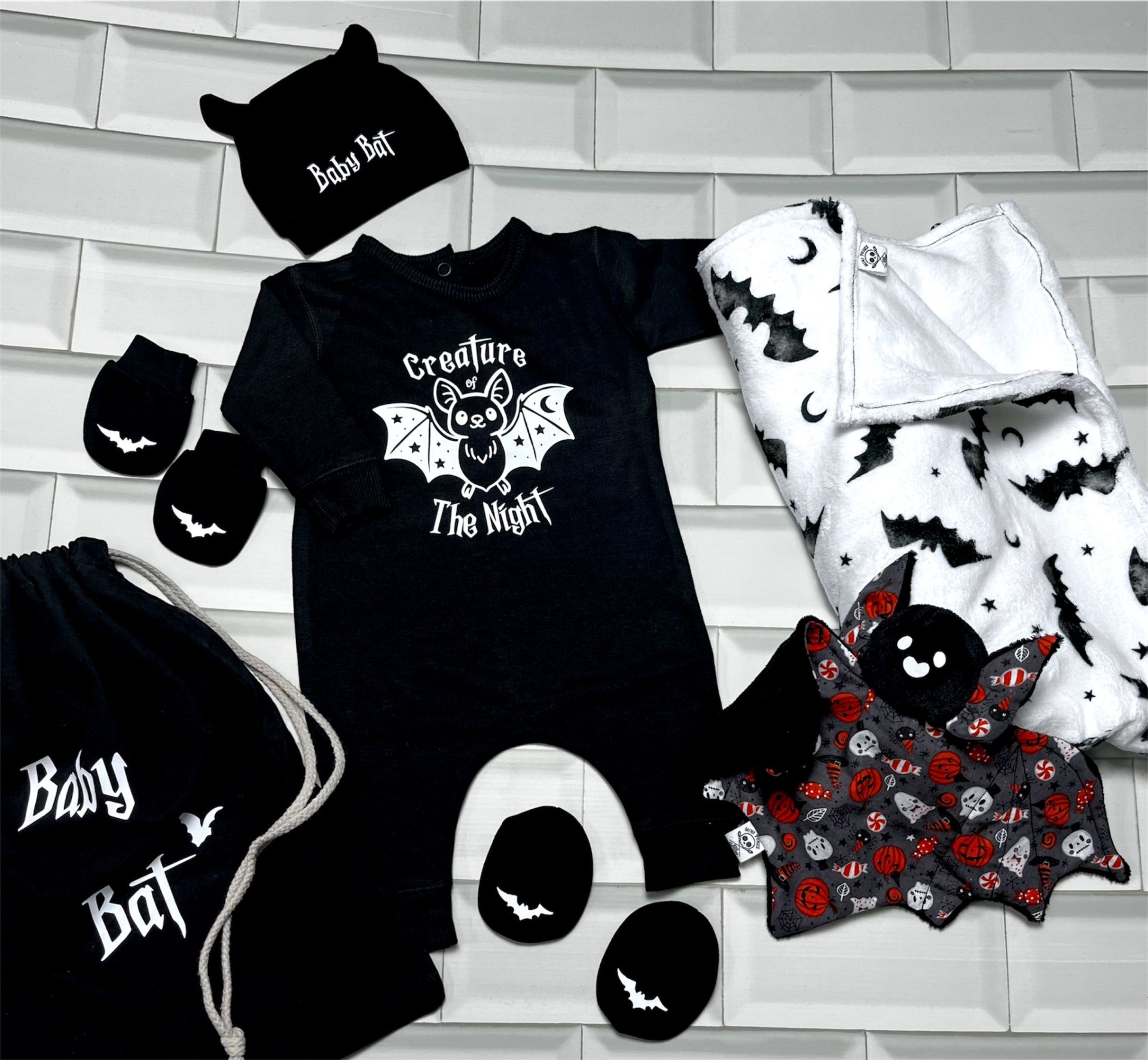 Conjunto de roupa e acessórios para bebé com tema morcego incluindo body, gorro, luvas, proteções, saco, manta e brinquedo.