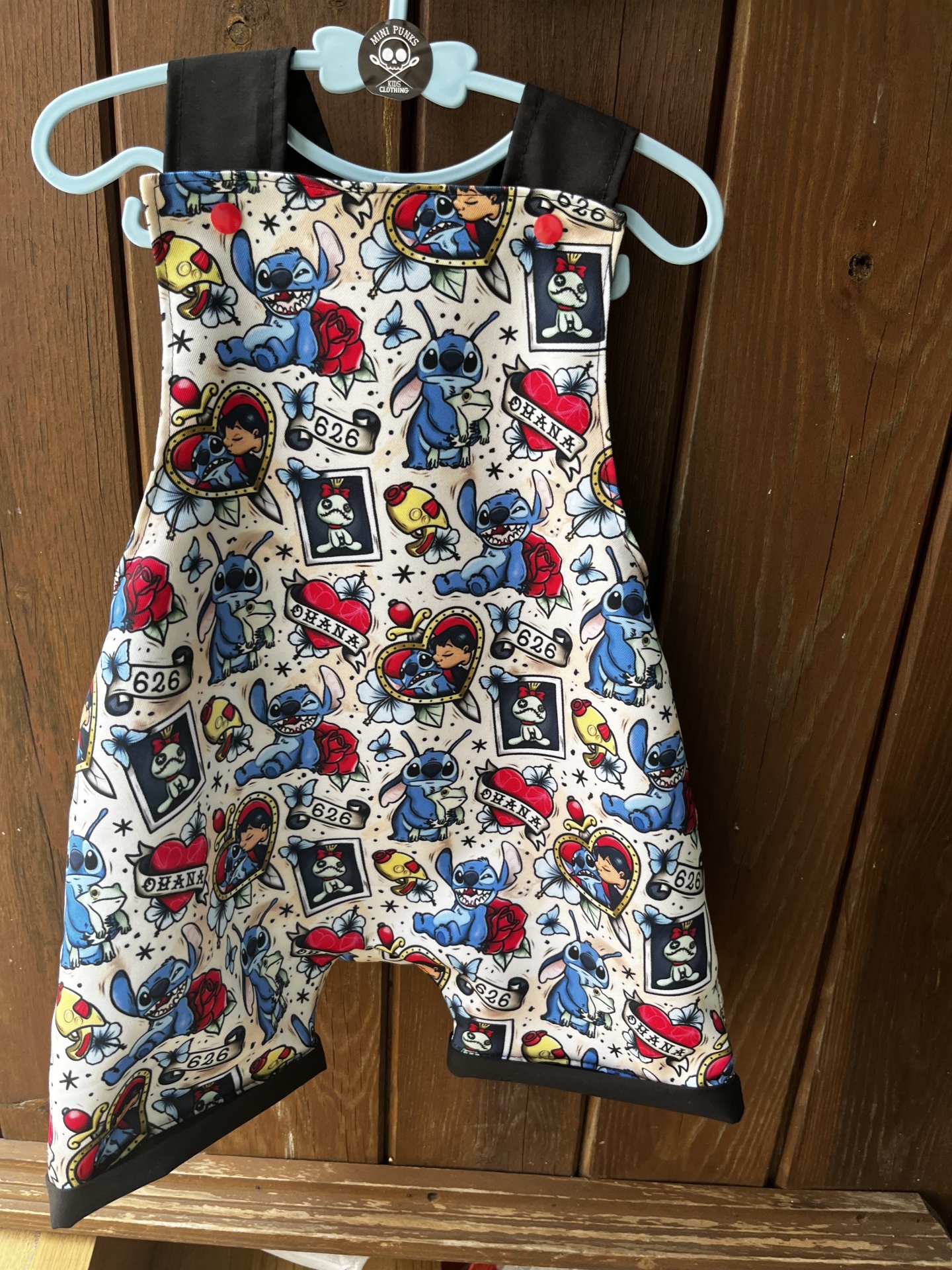 Macacão infantil com padrão de Stitch pendurado num cabide azul contra porta de madeira