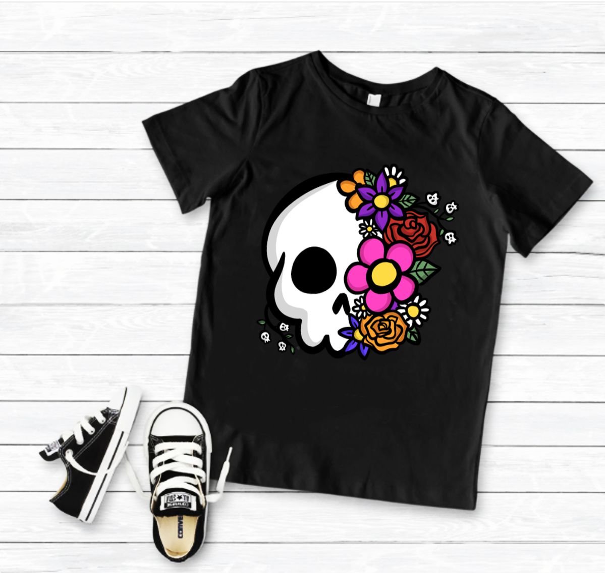 T-shirt preta com estampa de caveira e flores coloridas, com sapatilhas pretas e brancas ao lado sobre tábuas de madeira claras