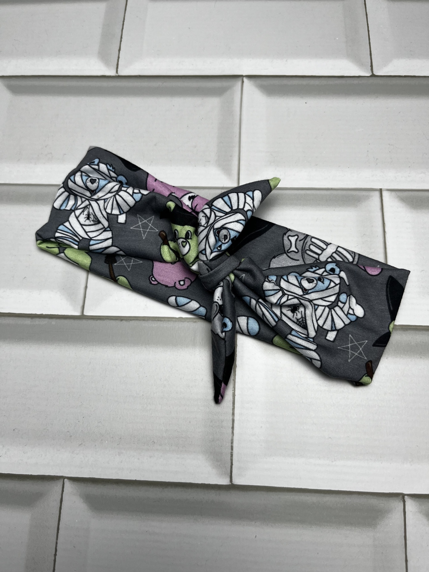 Faixa de cabelo preta com padrão de múmias e estrelas coloridas sobre azulejos brancos