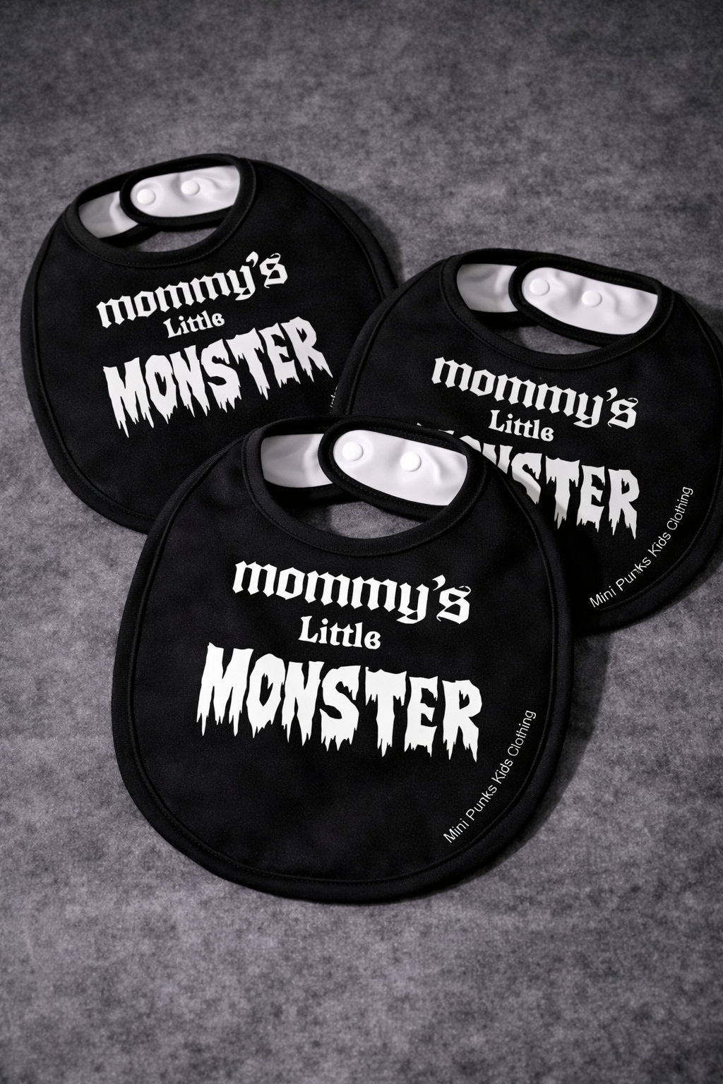 Três babetes pretos com texto branco colocados sobre superfície cinza.