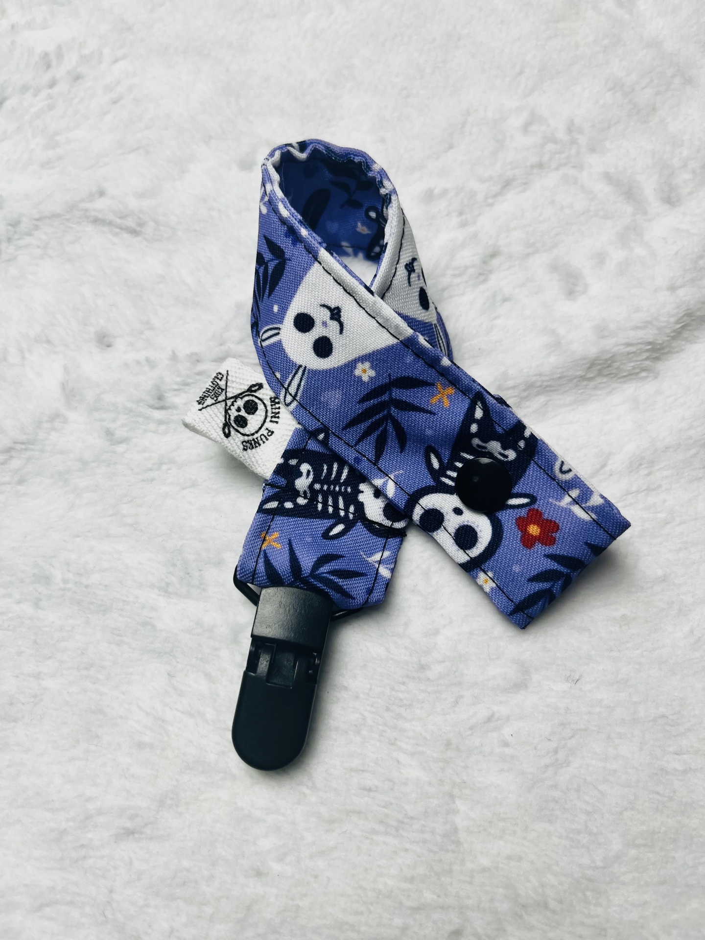 Tira azul com padrão de caveiras e flores com fecho de mola preto sobre fundo branco peludo.