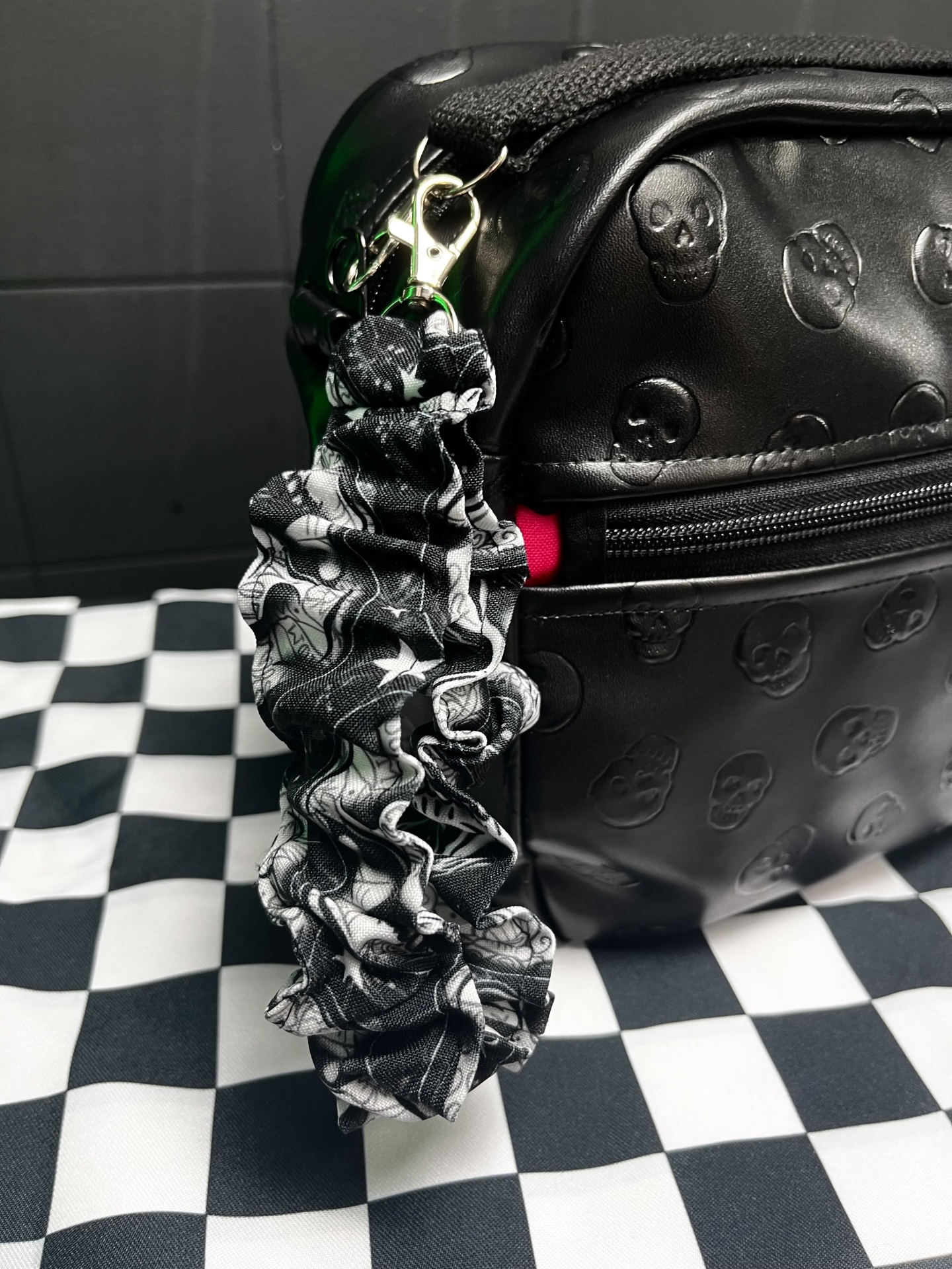 Mala preta com padrão de caveiras e acessório franzido de caveiras em fundo quadriculado preto e branco.