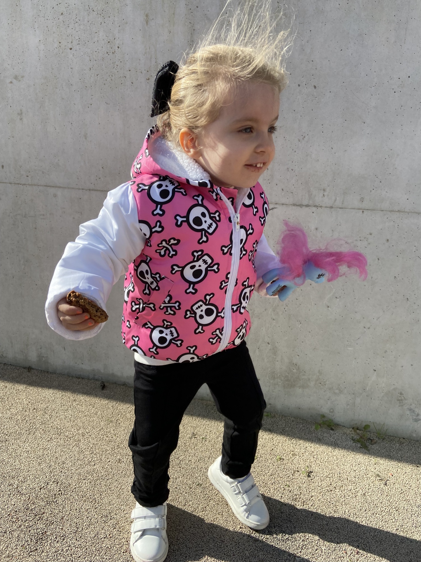 Criança com casaco rosa e branco, calças pretas e sapatilhas brancas segurando brinquedo colorido