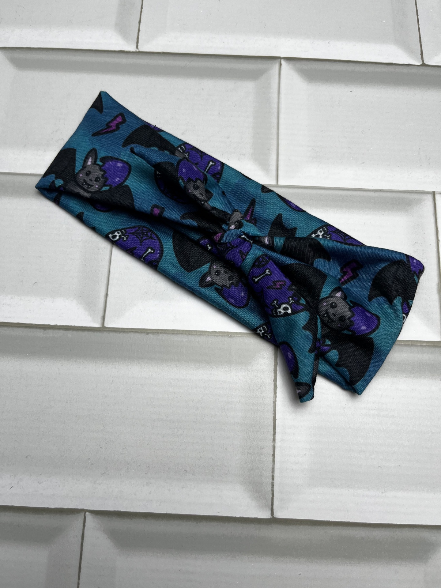 Faixa de cabelo azul escuro com padrão de morcegos e caveiras sobre azulejos brancos