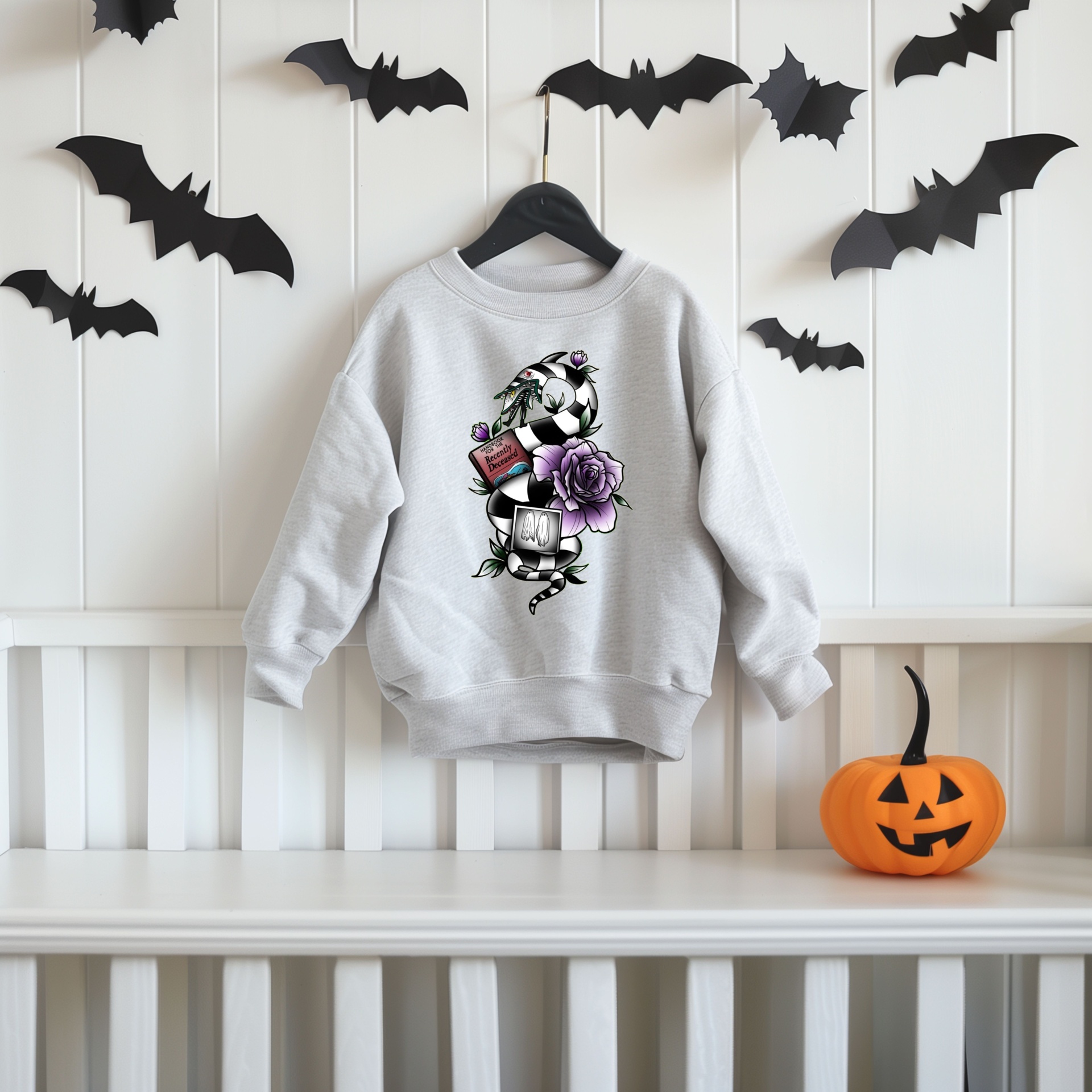 Camisola cinza clara com estampa de bola de futebol, flor roxa e garrafa, pendurada em cabide, com morcegos de papel e abóbora de Halloween.