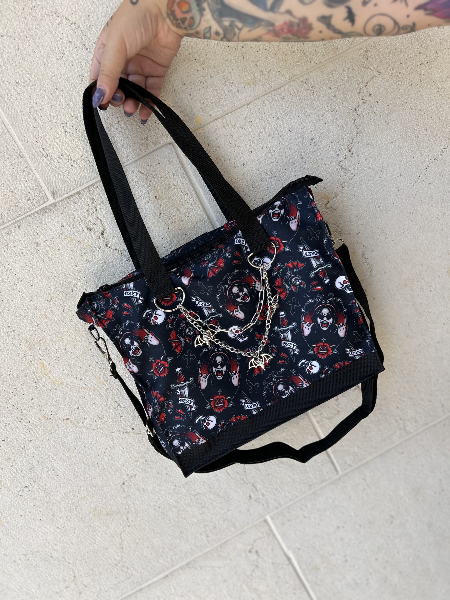 Mala preta com padrão colorido, correntes prateadas e alças pretas, segurada por mão tatuada.