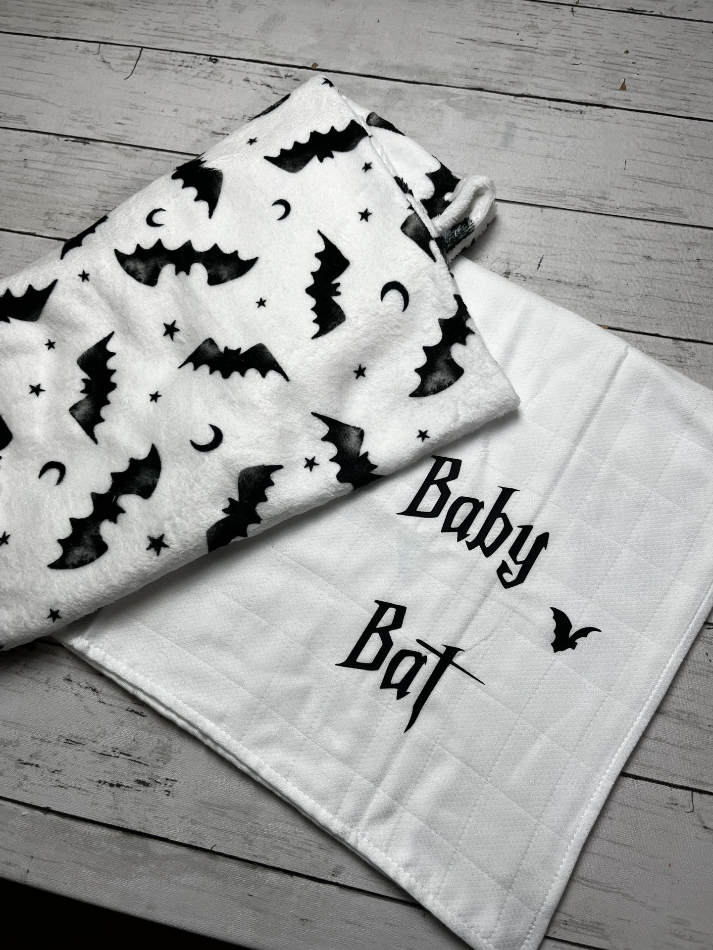 Dois panos brancos para bebé com padrões de morcegos e texto Baby Bat, sobre mesa de madeira clara.