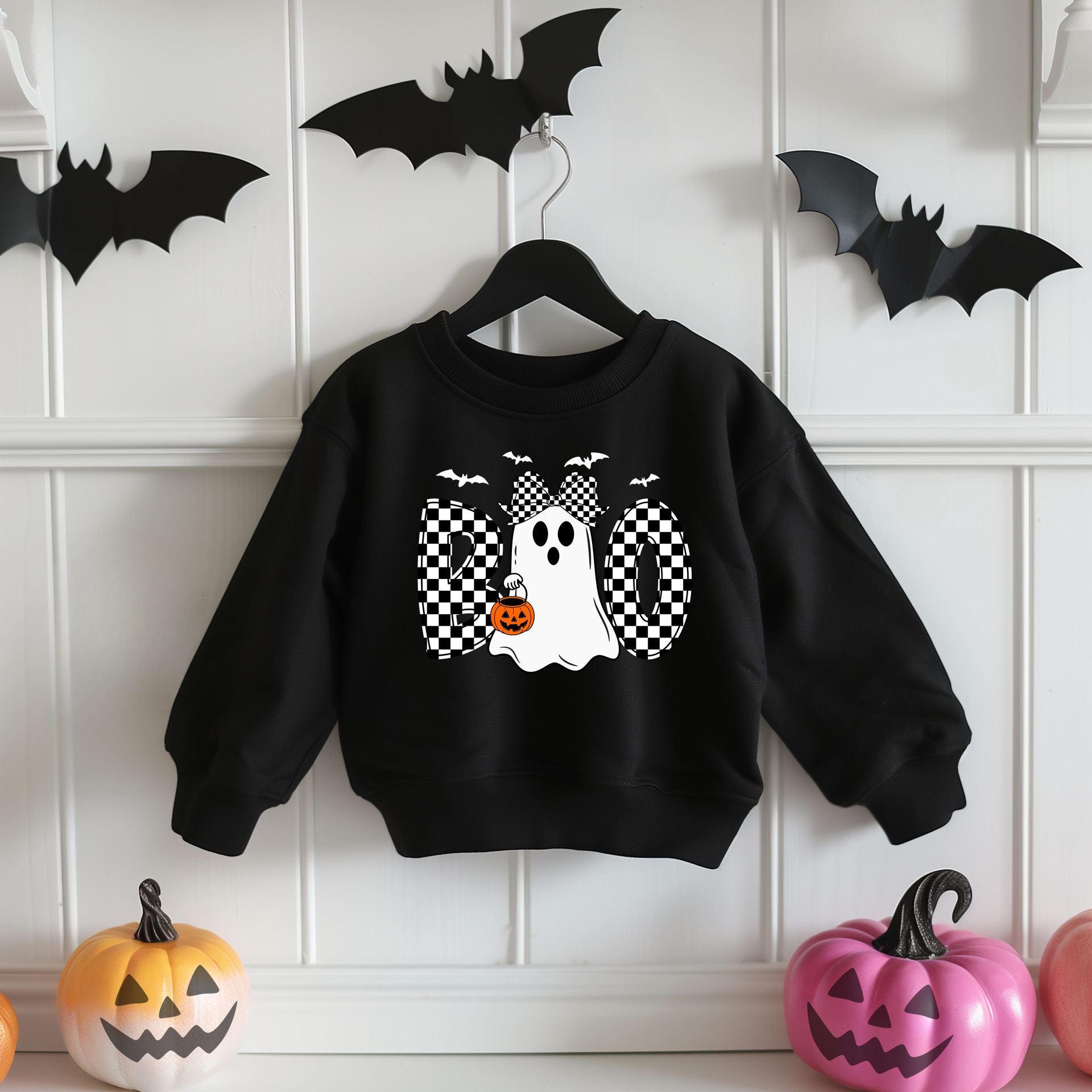 Sweatshirt preta com estampa de fantasma e letras BOO, pendurada em cabide contra parede branca com morcegos de papel e abóboras decorativas.