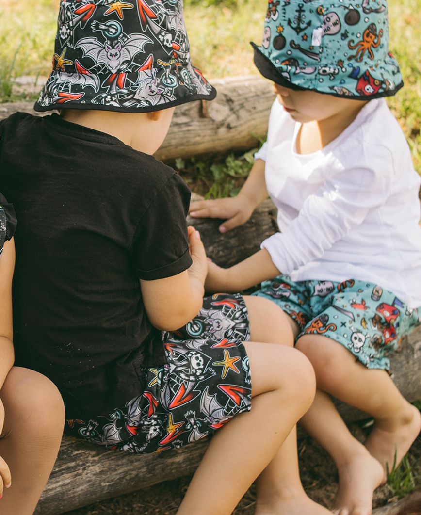 Duas crianças sentadas vestindo chapéus e calções com padrões temáticos coloridos, ao ar livre junto a um tronco de madeira.