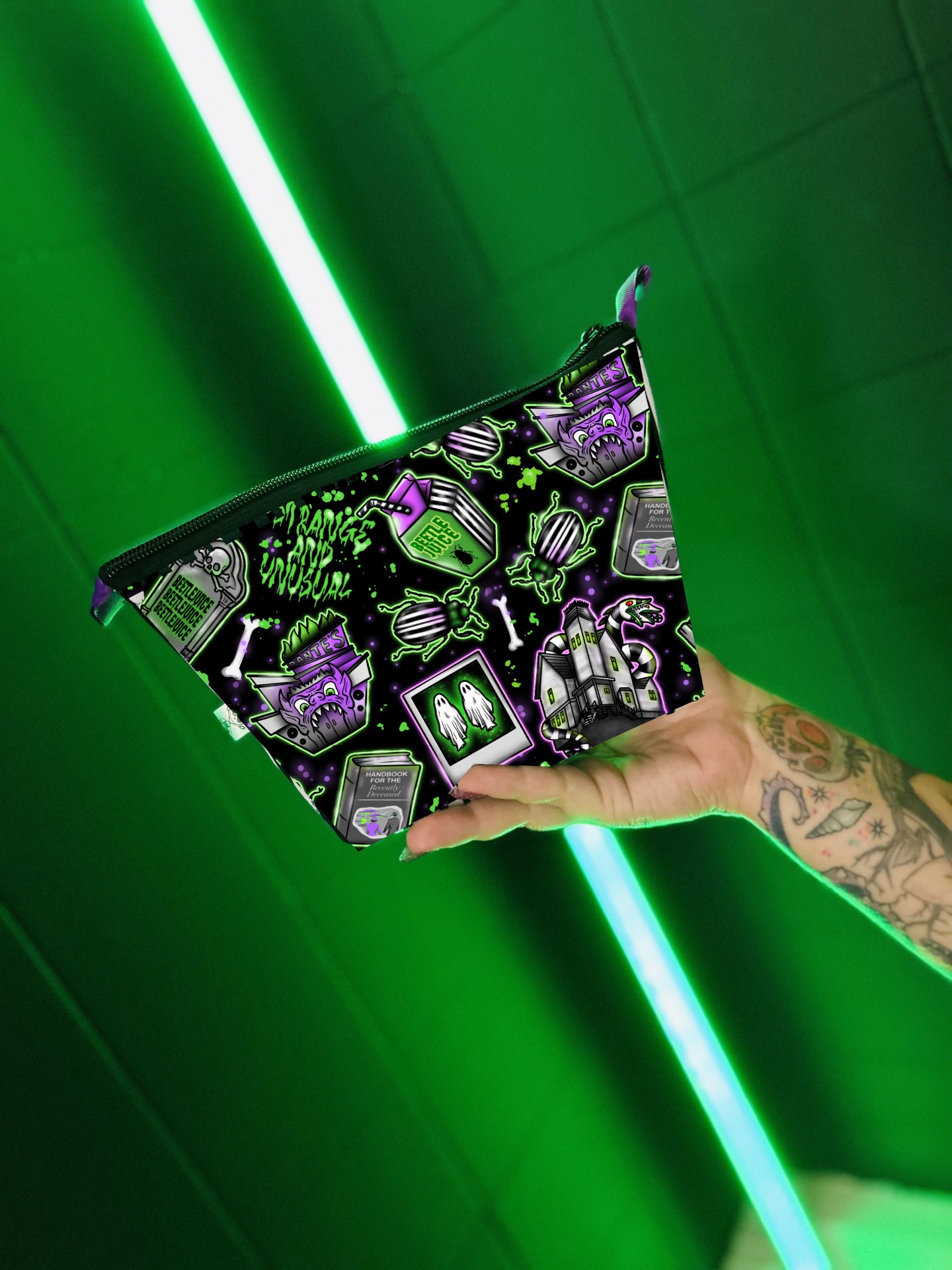 Necessaire preta com desenhos assustadores em roxo e verde, segurada por mão tatuada, fundo verde com luzes neon.
