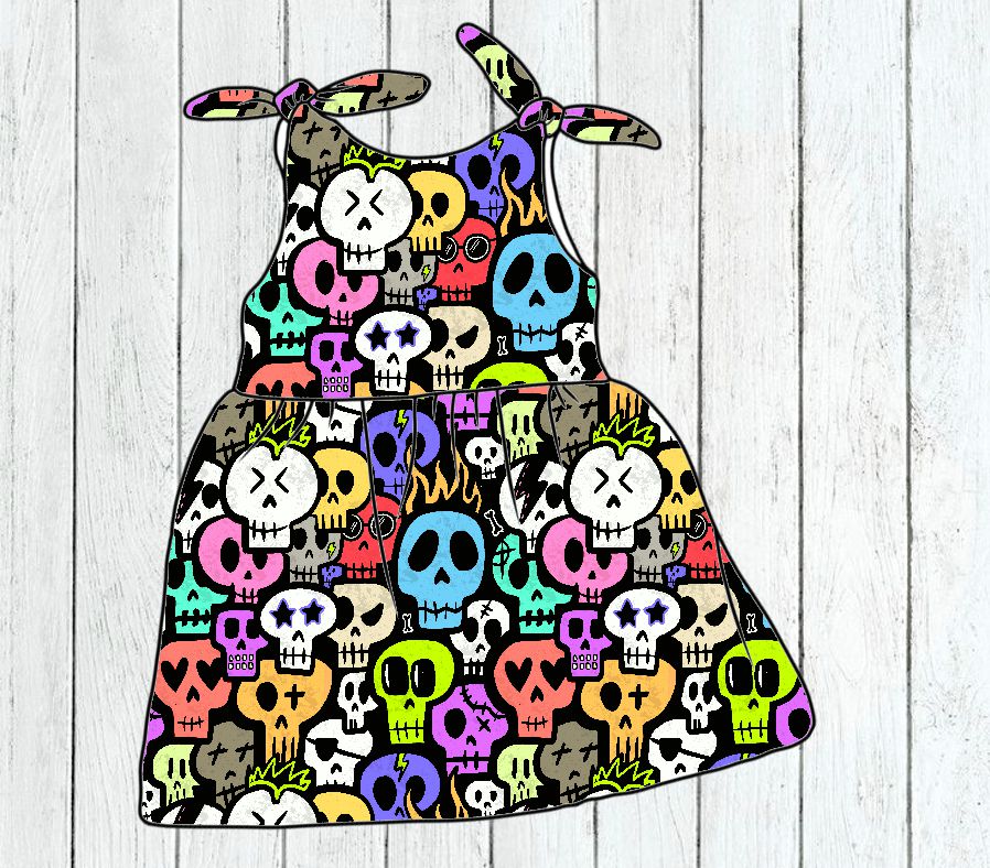 Vestido criança com padrão colorido de caveiras e tiras com laços em fundo de madeira clara.