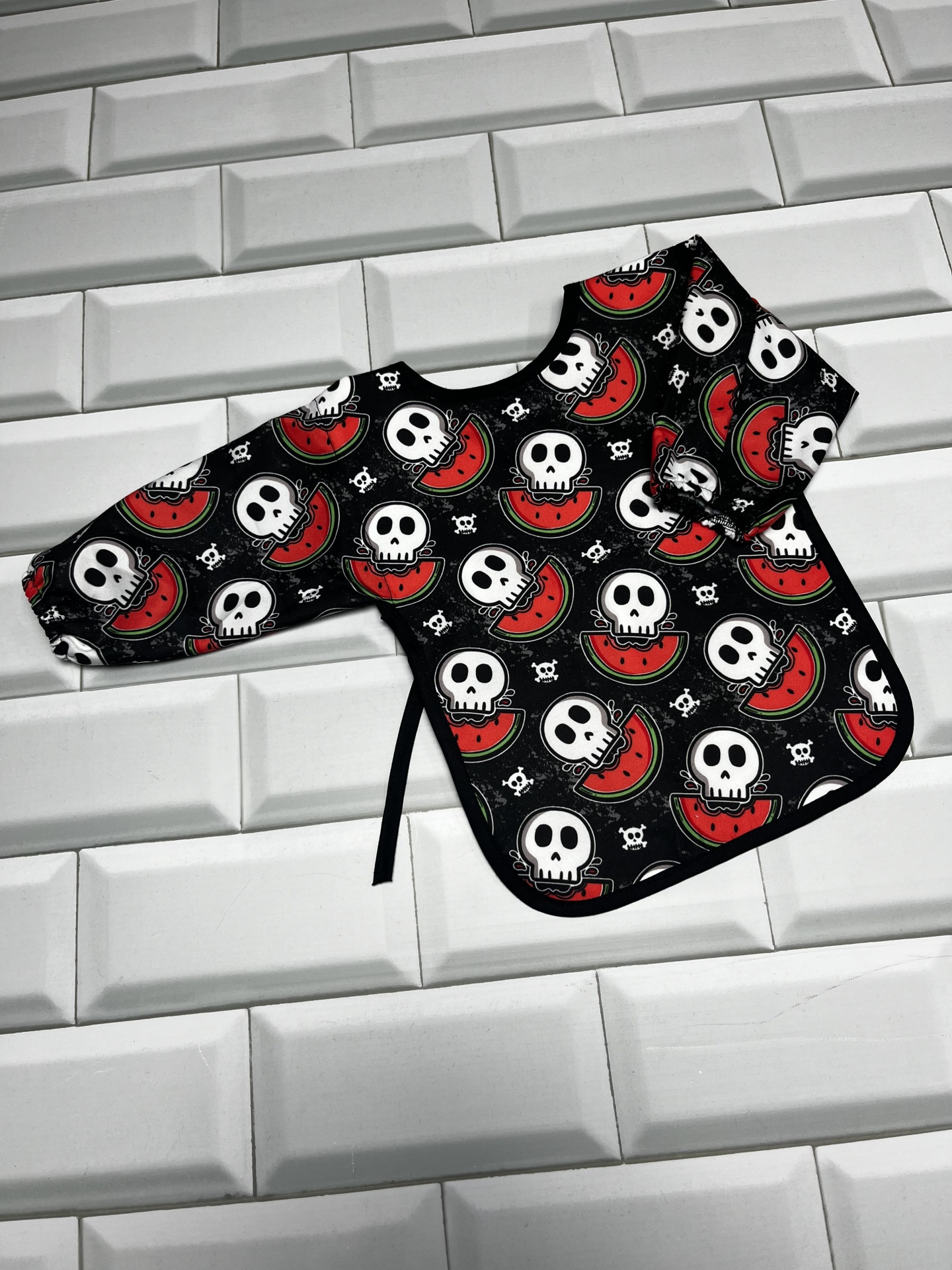 Babete de mangas compridas com padrão de caveiras e melancias sobre azulejos brancos