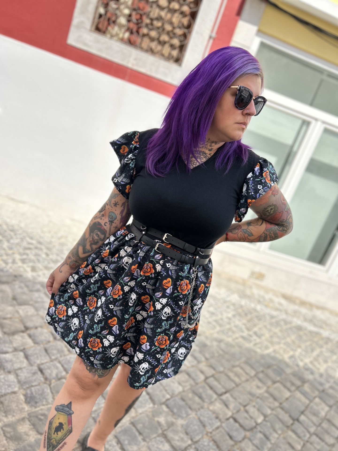 Vestido preto com padrão de caveiras e rosas, cinto preto, usado por mulher com cabelo roxo e óculos